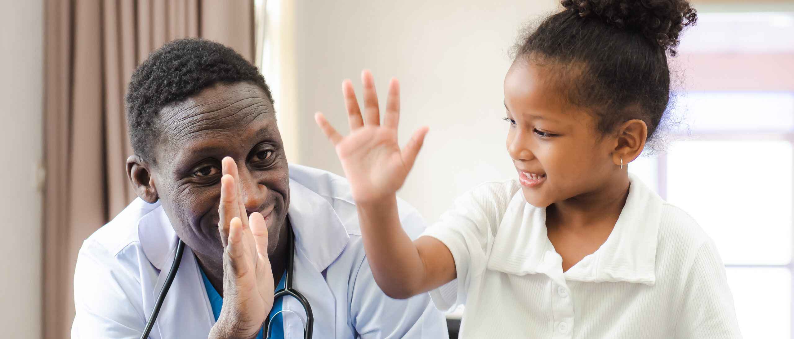 Child patient sitting in health center with African American doctor for medical care. Smiling little girl happy healthy after professional checkup at clinic. Practitioner, pediatrician checking, examining kid